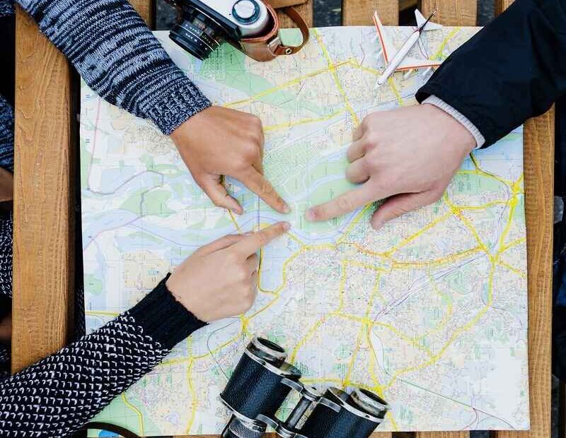 Three people pointing at a map while planning a trip, with a camera and binoculars on the table, symbolizing navigation and exploration using a map.